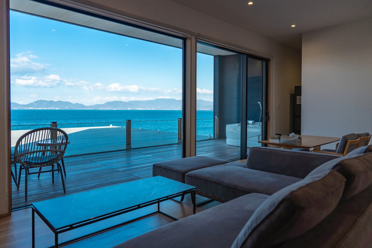 A modern living area is featured with large glass sliding doors that provide unobstructed views of the sea. A comfortable sectional sofa is positioned in front of a minimalist coffee table. A cozy chair sits nearby, enhancing the inviting space.