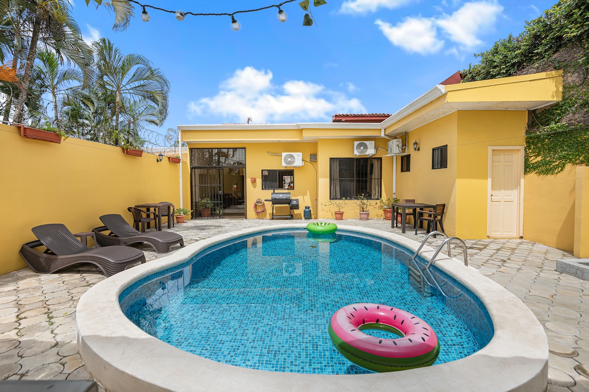 A private backyard is showcased with a clear blue pool, surrounded by lounge chairs. A green inflatable ring floats in the water, while tropical plants and string lights add a relaxed atmosphere. The villa's exterior features a bright yellow wall complemented by a partially visible door.