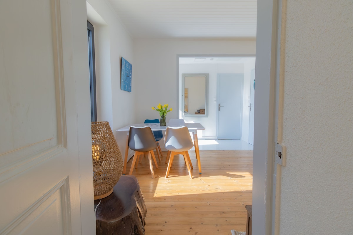 A well-lit dining area is visible through an open door, featuring a wooden table surrounded by four chairs. A vase with yellow flowers sits at the center of the table. Warm wooden flooring complements the light walls, enhancing the space's welcoming ambiance.