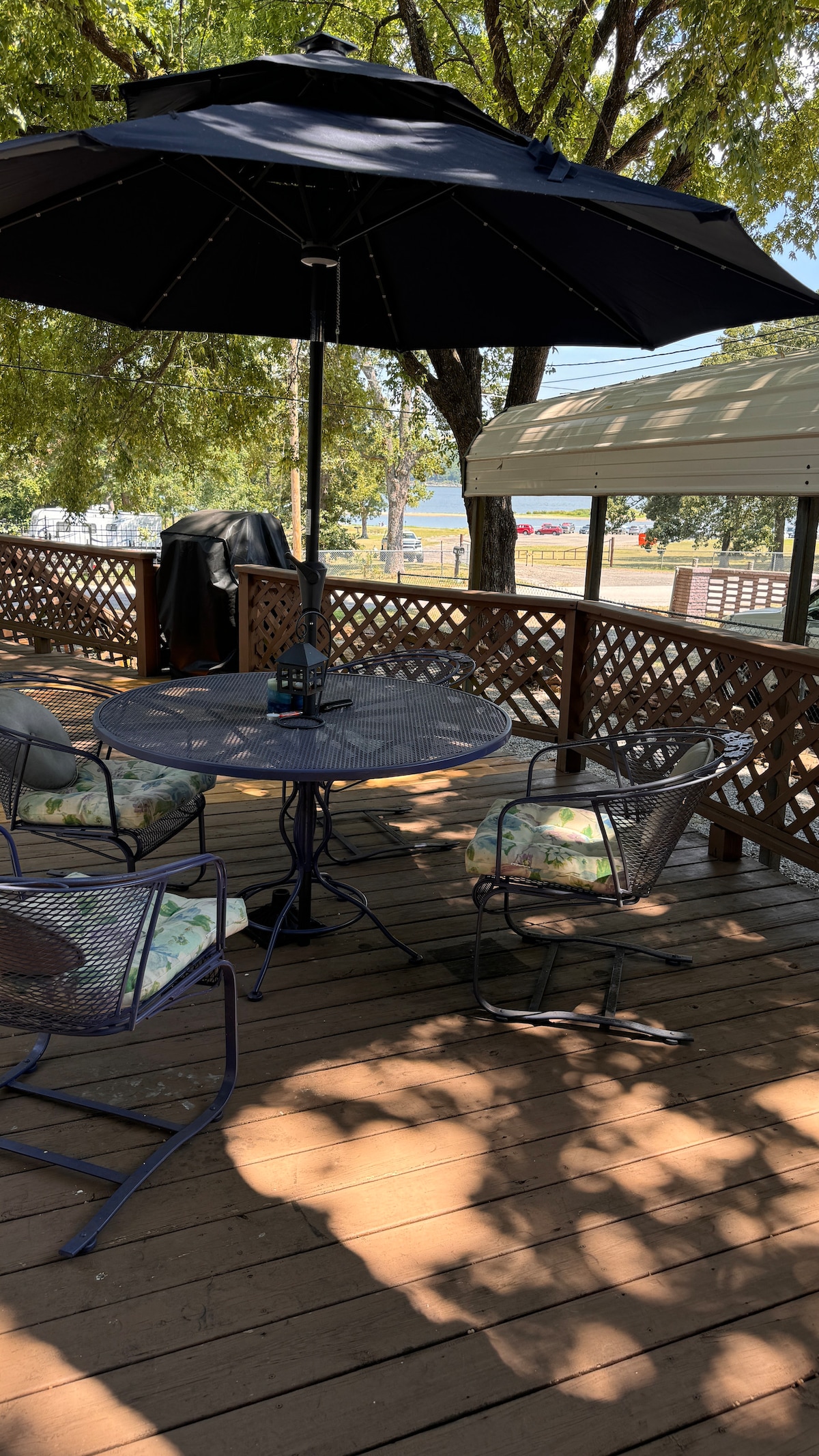 A spacious patio deck features a round table with chairs, topped by a large umbrella providing shade. The wooden flooring is adorned with dappled sunlight from overhanging trees. In the background, a glimpse of the lake can be seen, enhancing the outdoor dining experience.