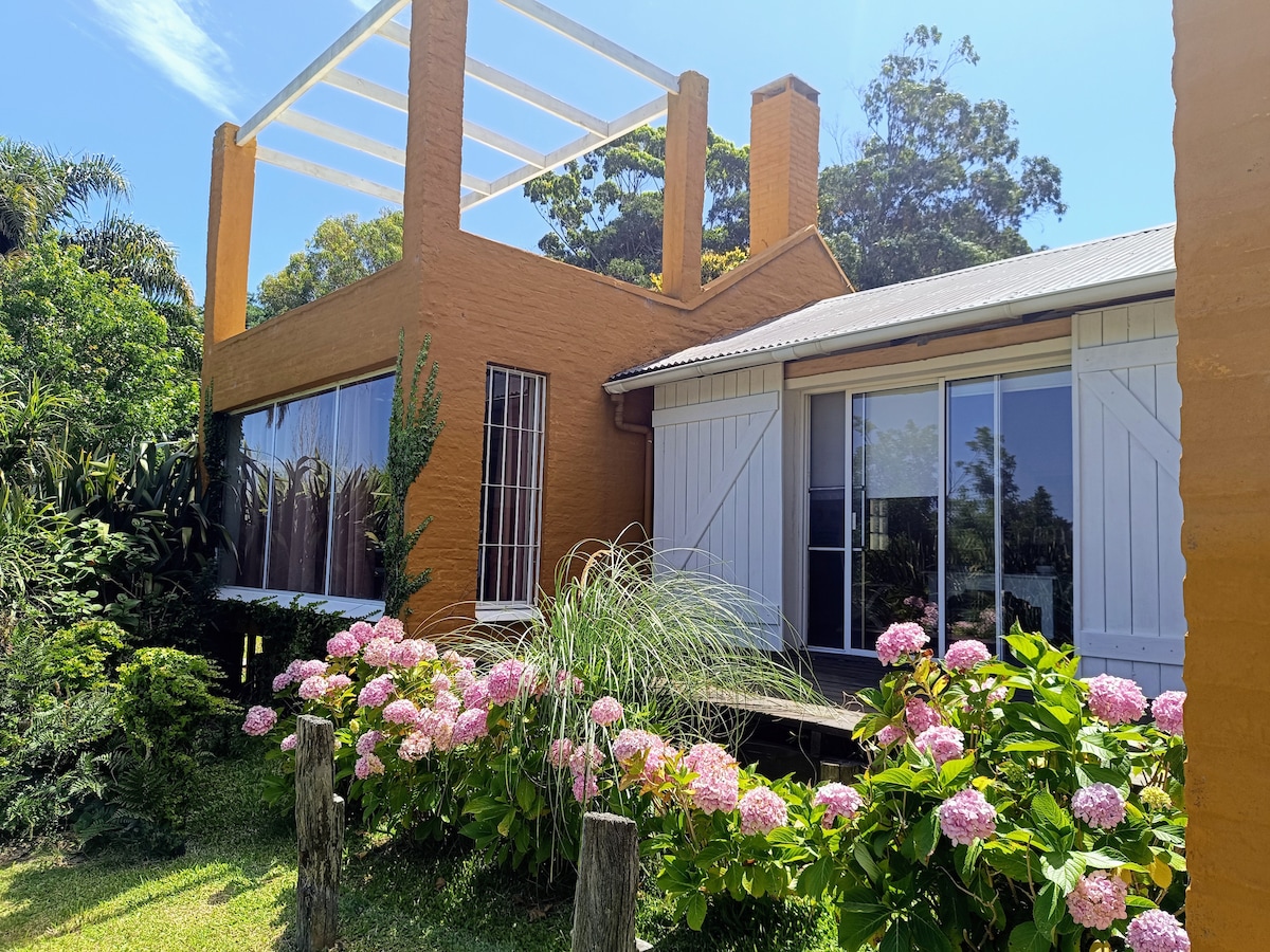 The exterior showcases a vibrant orange house framed by lush greenery and blooming hydrangeas. Large glass doors provide a view into the living space, while a partially open roof structure allows natural light to enhance the overall ambiance.