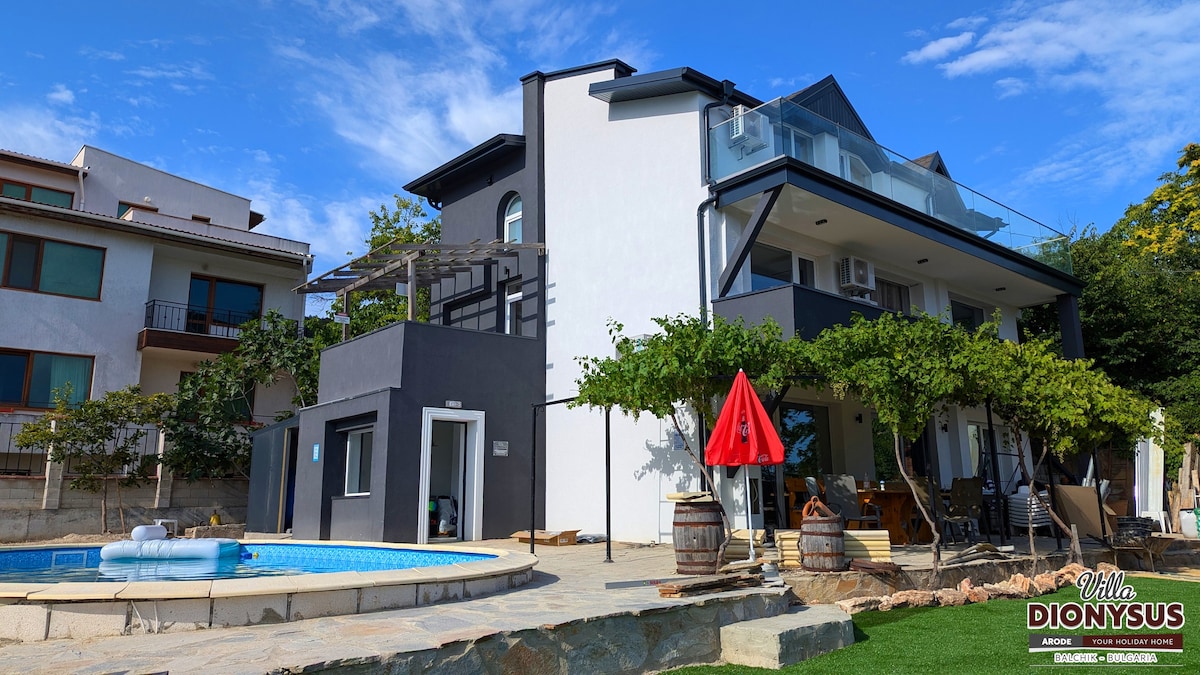 The villa features a modern, three-story structure with a swimming pool in the foreground. A red umbrella adds a pop of color to the outdoor seating area. Lush greenery surrounds the property, enhancing the inviting exterior, while the sky is clear with scattered clouds.