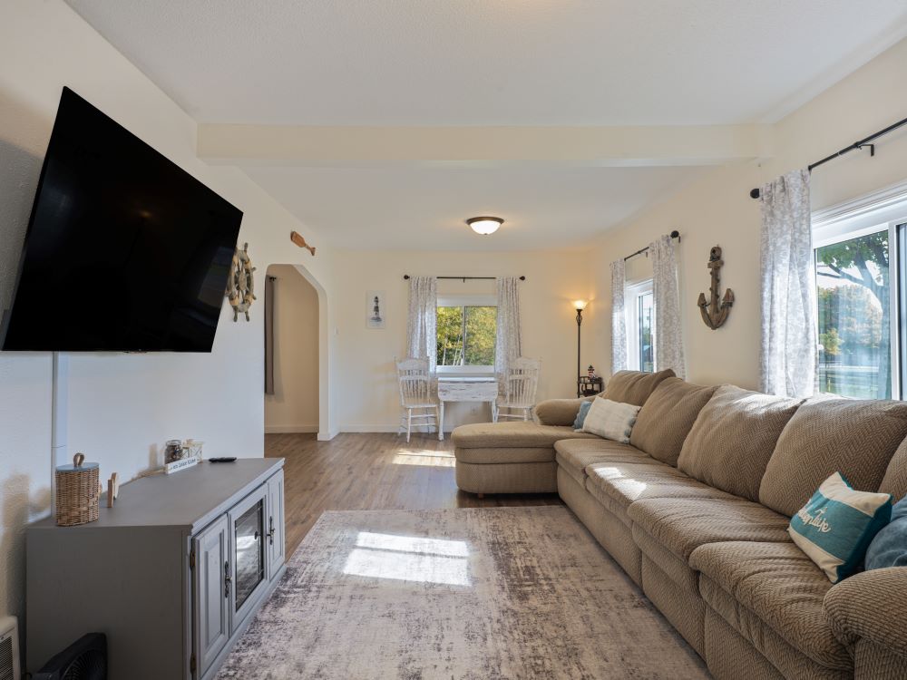 A spacious living area features a large sectional couch facing a flat-screen TV mounted on the wall. Natural light fills the room through wide windows, complementing a cozy dining area set for four. Subtle decor enhances the relaxed atmosphere of the space.