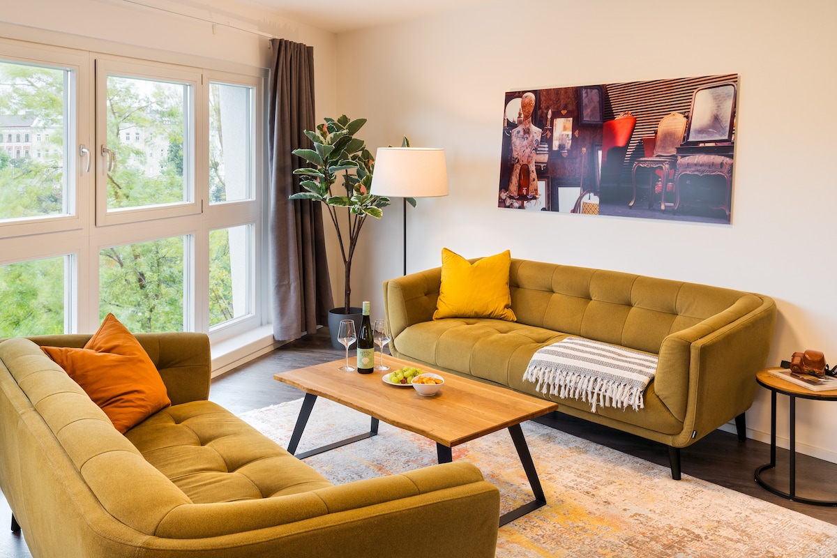 A living area is presented featuring two green sofas arranged around a wooden coffee table. A floor lamp provides ambient lighting, while a decorative plant adds a touch of nature. A large window lets in natural light, framed by gray curtains, and wall art enhances the space.
