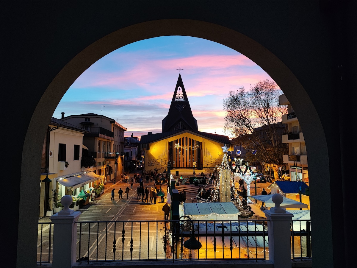 A view is captured through an archway, showcasing a vibrant square at sunset. Stalls and festive decorations line the square, with people walking about. The silhouette of a distinctive church spire is visible against the colorful sky, creating a warm atmosphere.