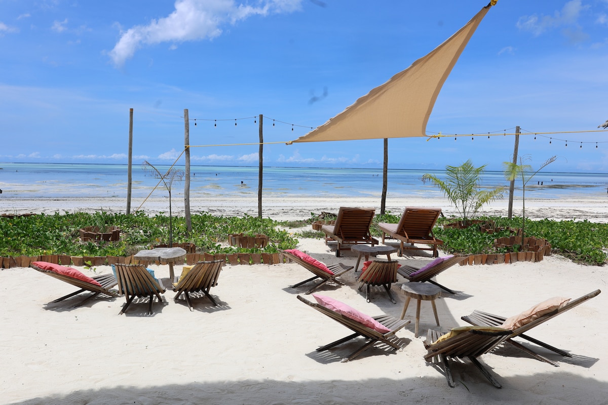 A sandy area on the beach is furnished with several wooden loungers, some adorned with colorful cushions. A large fabric shade is positioned overhead, providing shelter from the sun. The calm sea and clear sky extend into the distance, enhancing the serene coastal setting.