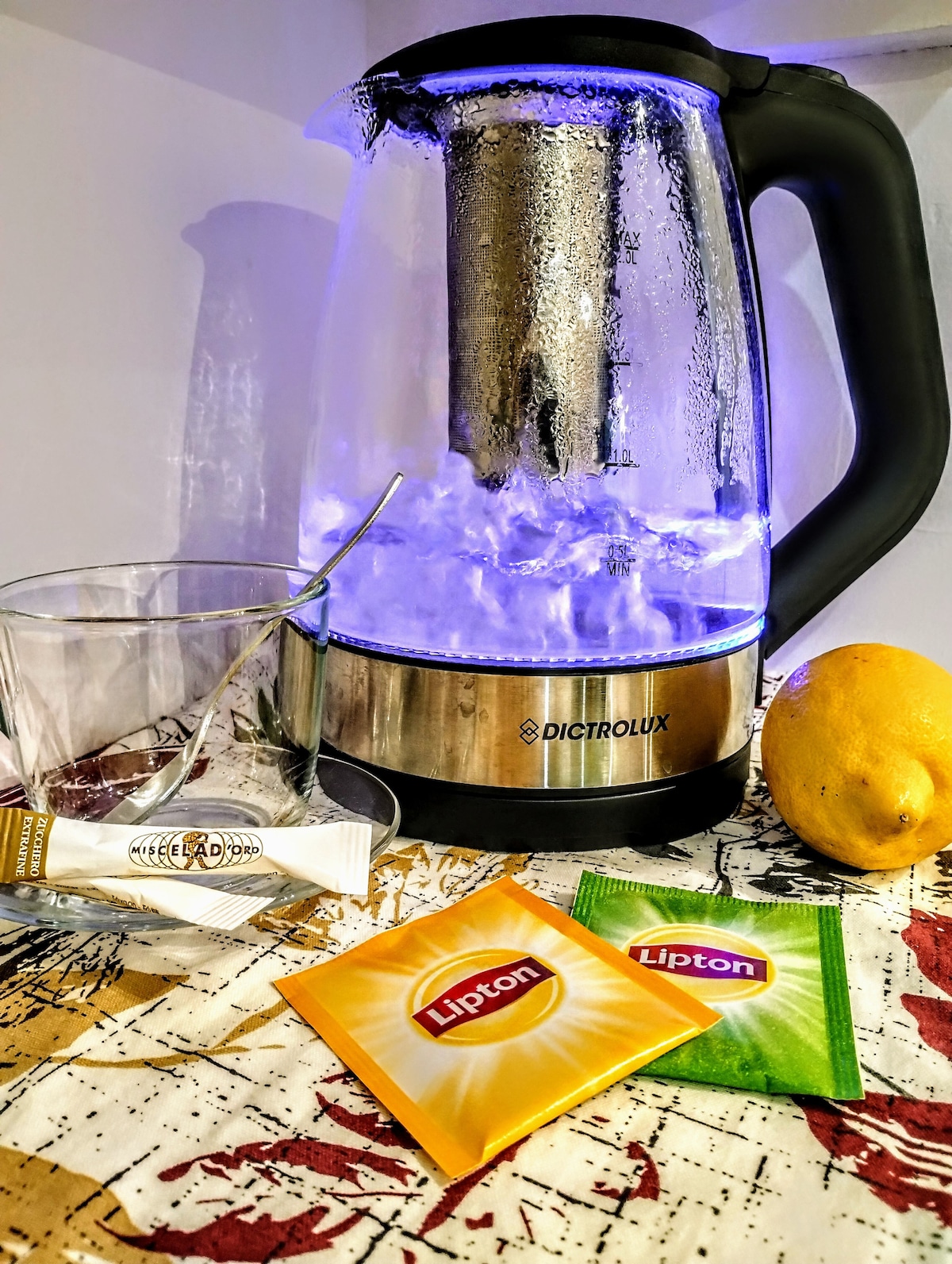 An electric kettle with a transparent body is seen boiling water, revealing a stainless steel interior. A glass is positioned beside the kettle, along with tea sachets in yellow and green. A lemon rests next to the kettle on a patterned surface, enhancing the inviting setting.