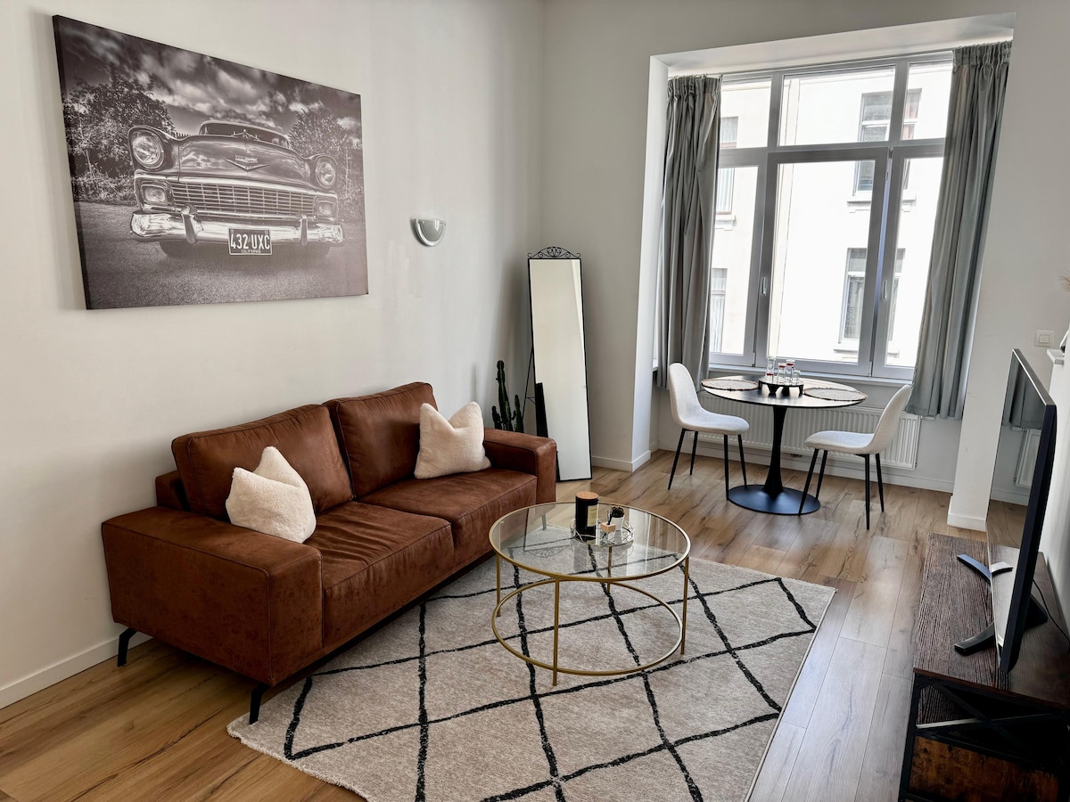 A modern living area features a brown sofa adorned with white pillows, complemented by a round glass coffee table atop a patterned area rug. A large mirror is positioned beside a bright window that invites natural light, while a dining table for two is visible in the background.