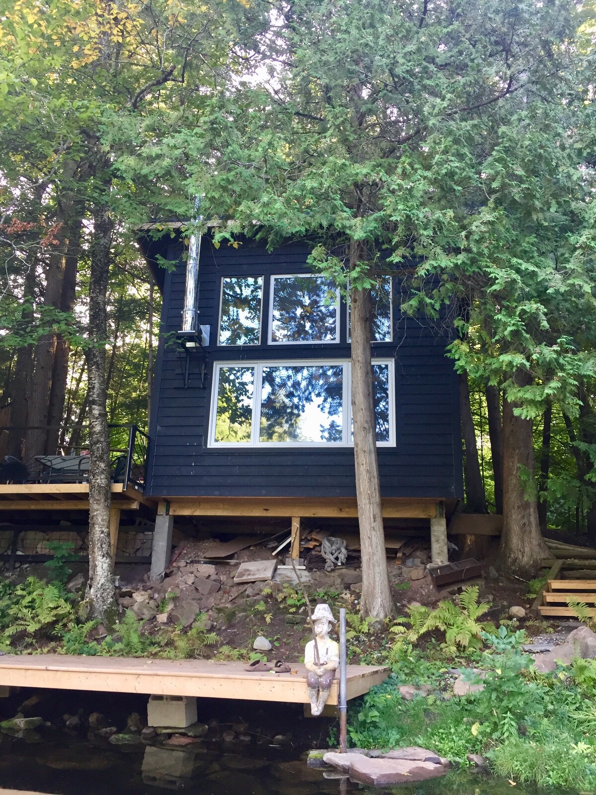 A modern timber-frame cabin is situated on stilts among trees, overlooking Nogies Creek. Its large windows reflect natural light while providing views of the surrounding greenery. A small wooden dock leads to the water, enhancing the serene outdoor experience.