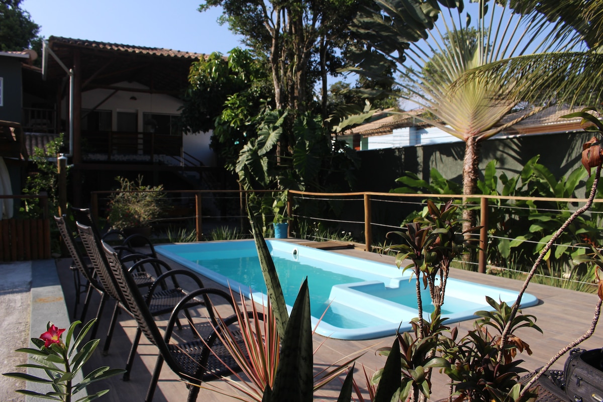 A calming outdoor space features a glistening pool surrounded by lush greenery and several black lounge chairs. Tropical plants frame the area, enhancing the serene ambiance. The setting is complemented by a wooden deck that invites relaxation and peaceful moments.