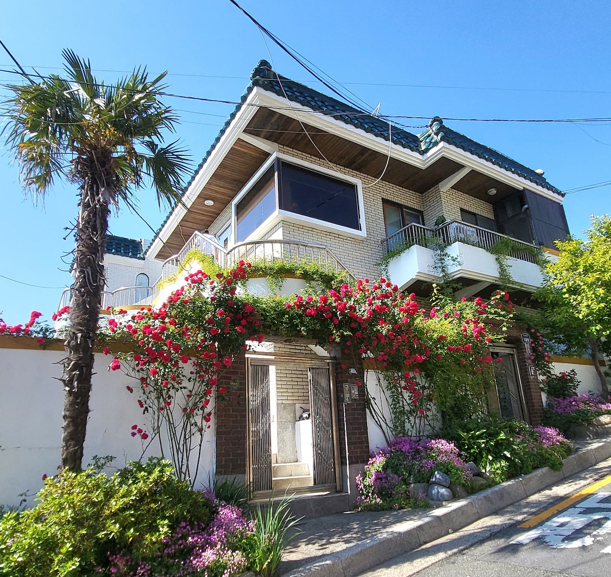 A charming two-story building is surrounded by vibrant flowers and greenery. The facade features enchanting floral arrangements draping from the balcony. A clear blue sky complements the structure, which has a tiled roof and large windows that enhance natural lighting.