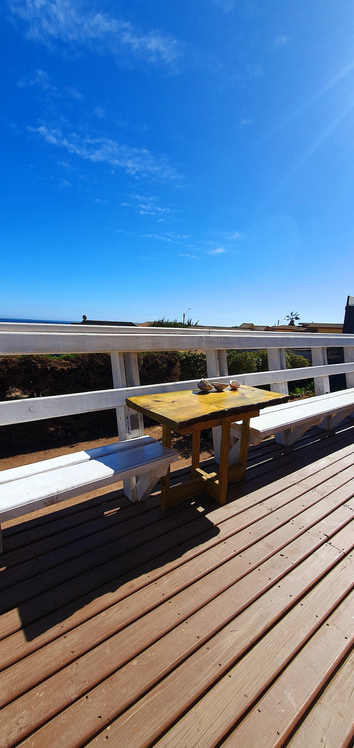 A bright terrace features a wooden table placed beside a bench. The blue sky overhead enhances the open atmosphere, while a view of the surrounding greenery is visible beyond the railing. The space invites relaxation in a scenic outdoor setting.
