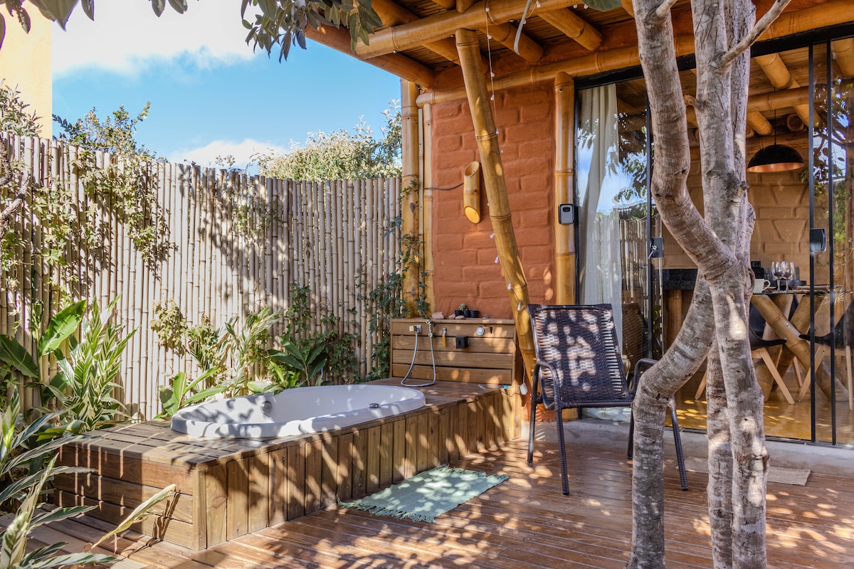 An inviting deck features a warm wooden surface surrounding a private hot tub, with lush greenery framing the space. Bamboo fencing offers seclusion, while a comfortable chair is positioned nearby, and a portion of the structure is visible in the background.