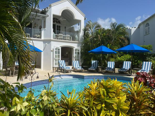 A serene outdoor area features a private pool surrounded by lush tropical plants. Seating areas with blue and white striped lounge chairs are shaded by blue umbrellas. The villa's elegant architecture is complemented by large glass doors that open to the inviting pool.