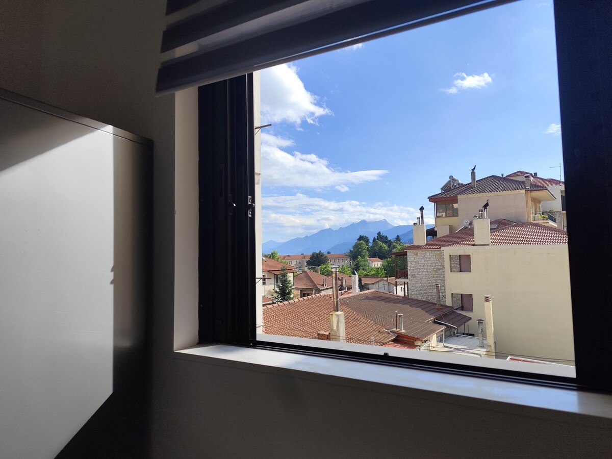 A view from the window showcases a landscape of rooftops with a backdrop of distant mountains. The bright blue sky features scattered clouds, providing a sense of openness and tranquility. Light filters through the window, illuminating the interior space.