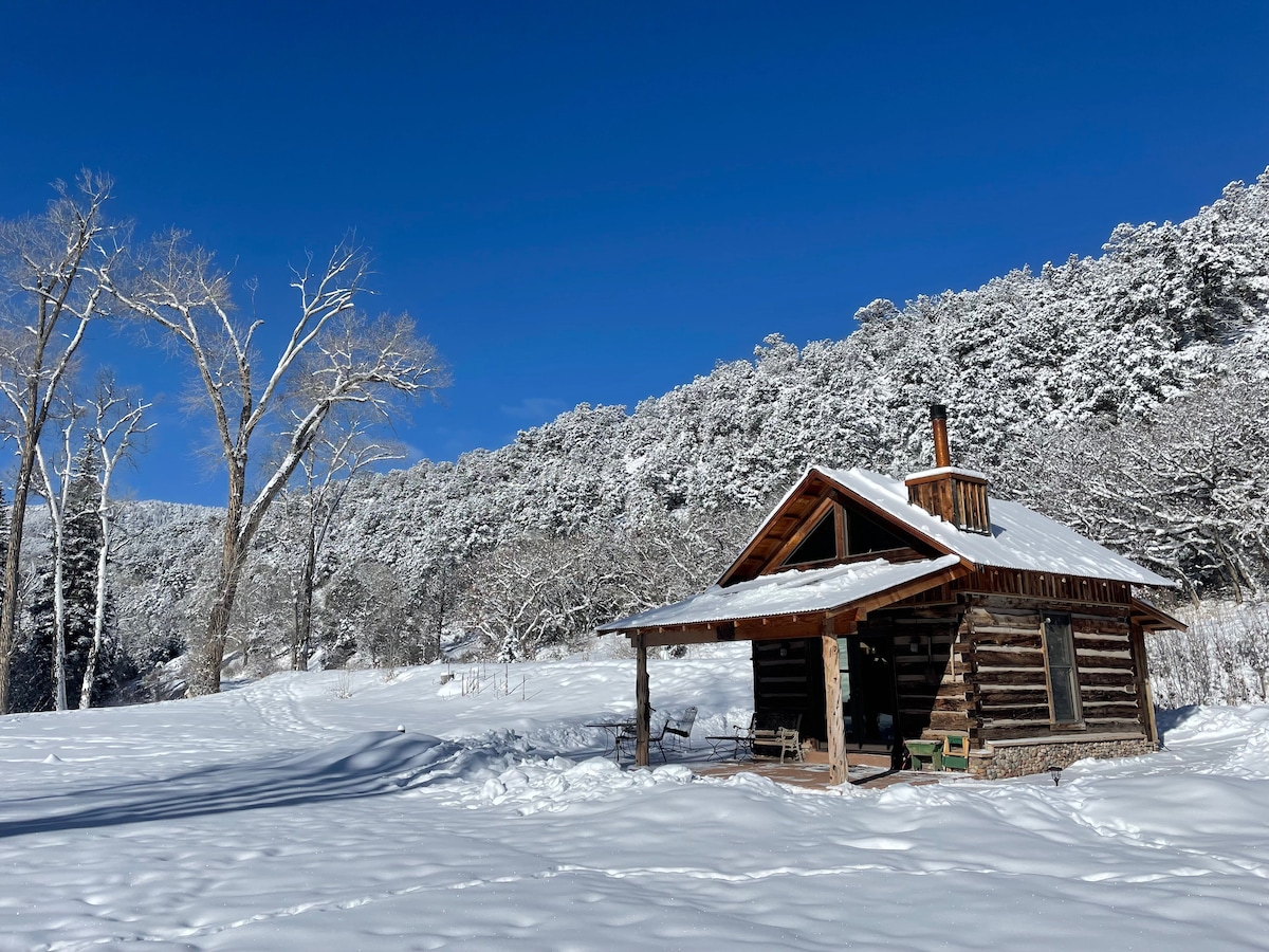 A remote cabin is nestled in a snowy landscape, surrounded by tall trees and rolling hills. The rustic structure features a sloped roof, a chimney, and a welcoming porch with chairs, all blanketed under fresh snow against a clear blue sky.