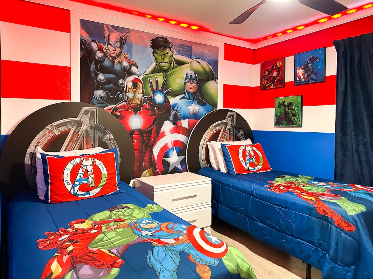 Მოირგეთ შურისმაძიებლების ამ საძინებელში: გამუქებული წითელი და თეთრი ზოლები ხვდება ღრმა ლურჯ კედლებს, ჩარჩოში მოქცეული ტყუპი საწოლები, რომლებიც გამოწყობილია ძლიერი სუპერგმირის თეთრეულში და შესაბამისი ბალიშები. Ცენტრალური ტუმბო, ჭერის ვენტილატორი კაპიტანი ამერიკა ფართან ერთად               