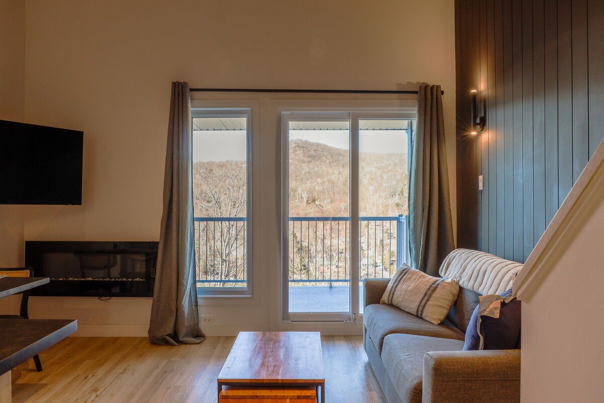 A cozy living area features a comfortable sofa draped with decorative cushions. Large sliding glass doors open to a balcony, providing a view of the surrounding mountains. A wall-mounted television is positioned for easy viewing, and natural light fills the space.