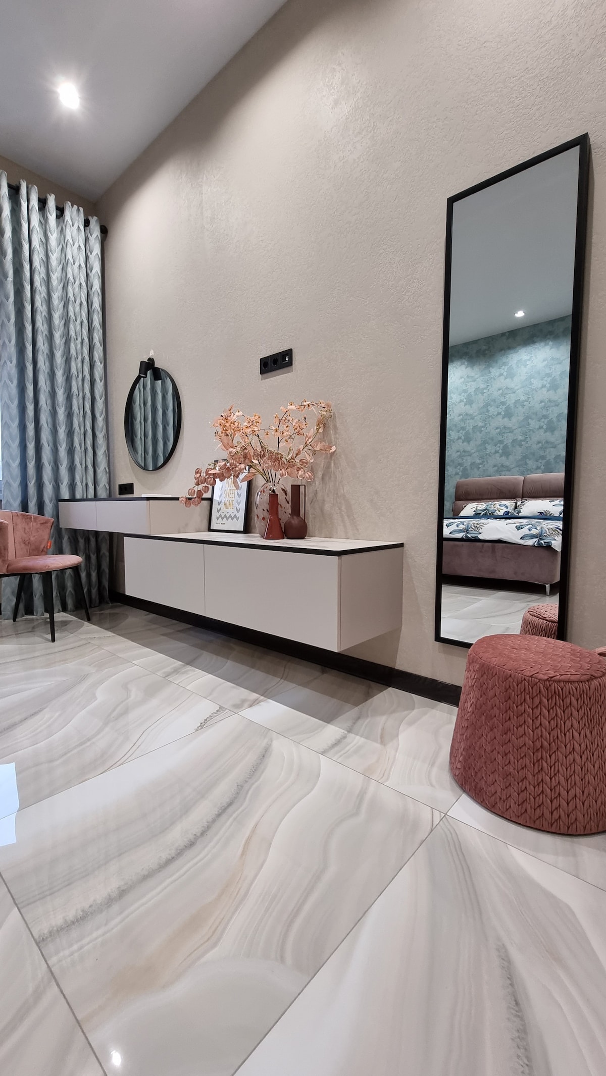 A well-designed corner features a low cabinet adorned with decorative items, including a vase of branches. A full-length mirror reflects the space, while soft textures are introduced through a plush chair and round pouf. Light-colored, glossy tiles enhance the overall brightness.