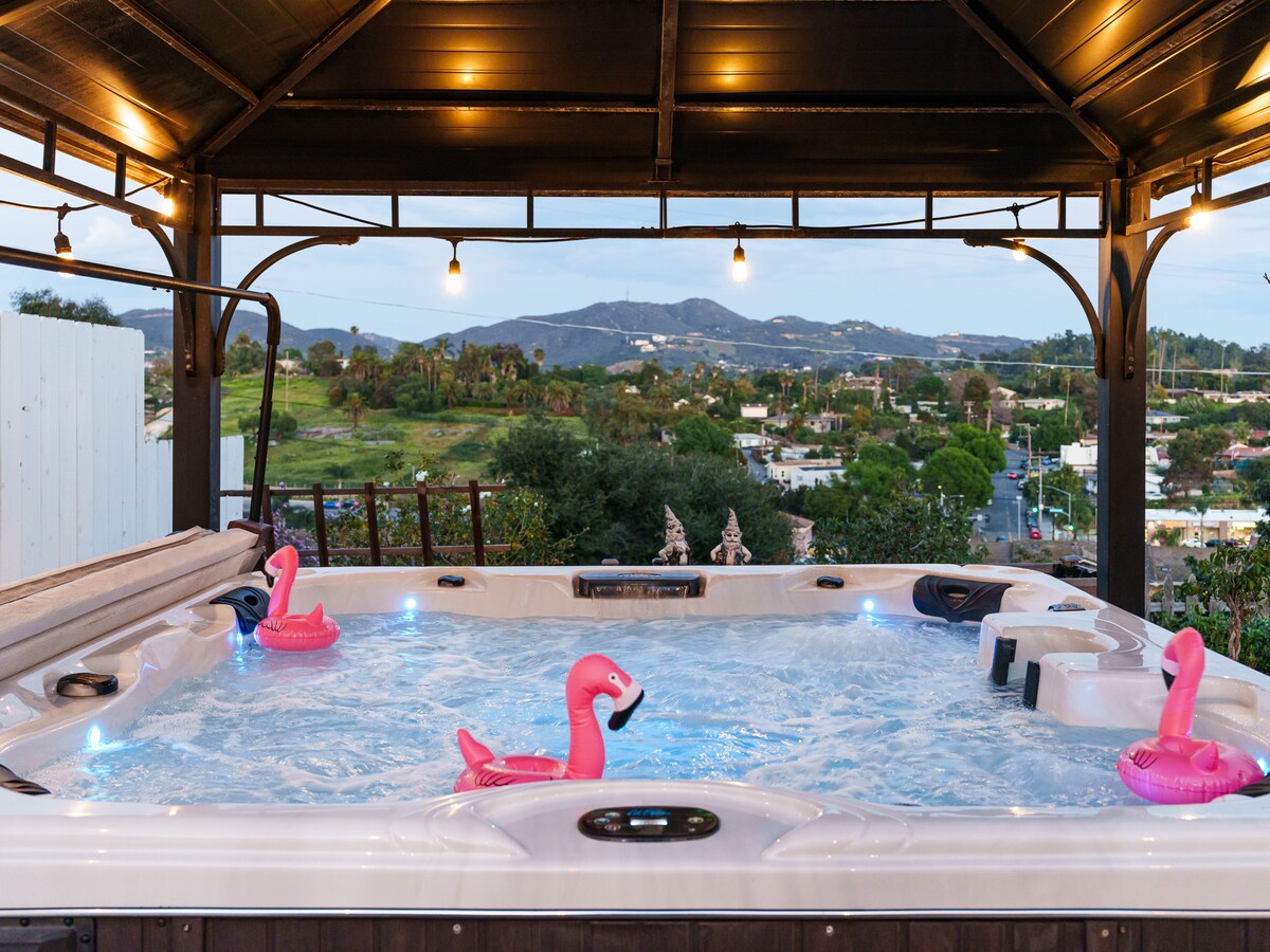 A hot tub is positioned under a roofed gazebo, surrounded by tranquil mountain views. Soft lighting enhances the atmosphere, while colorful flamingo floaties are seen resting in the water. The expansive view of the surrounding landscape creates a serene setting for relaxation.