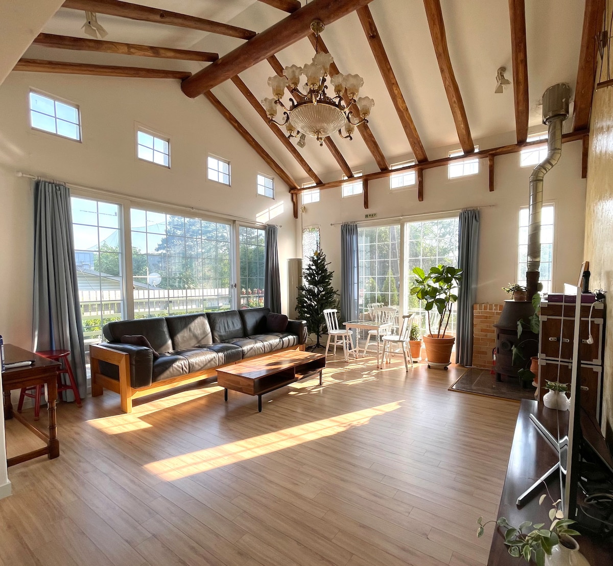 A spacious living area features large windows that flood the space with natural light, highlighting the wooden beams above. A comfortable leather sofa is positioned alongside a coffee table, with potted plants adding a touch of greenery. A decorative chandelier hangs elegantly from the ceiling.