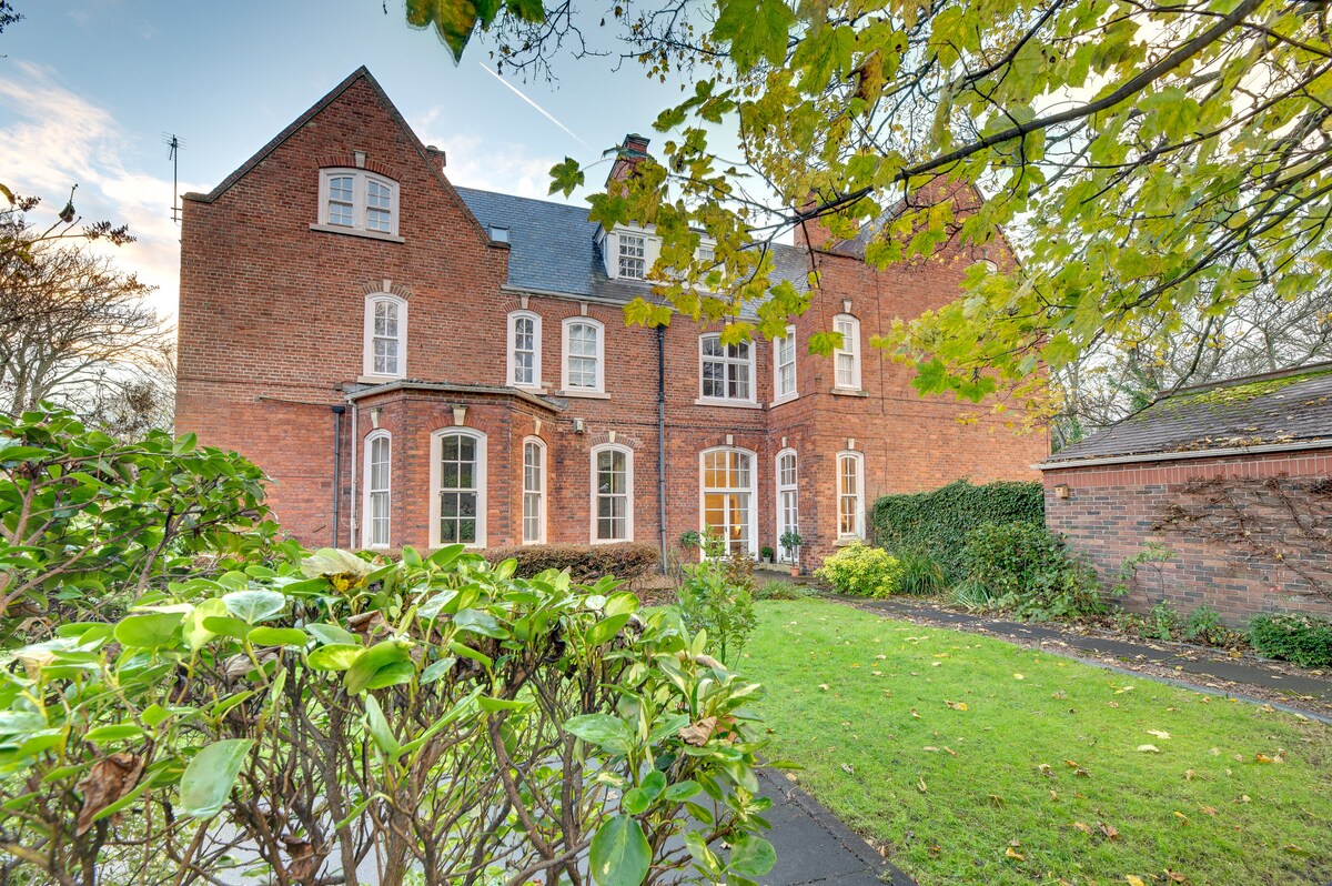 The exterior of a historic brick building showcases architectural features and large windows, surrounded by lush greenery and a manicured lawn. A pathway leads to the entrance, framed by foliage and trees, creating a peaceful and inviting atmosphere.