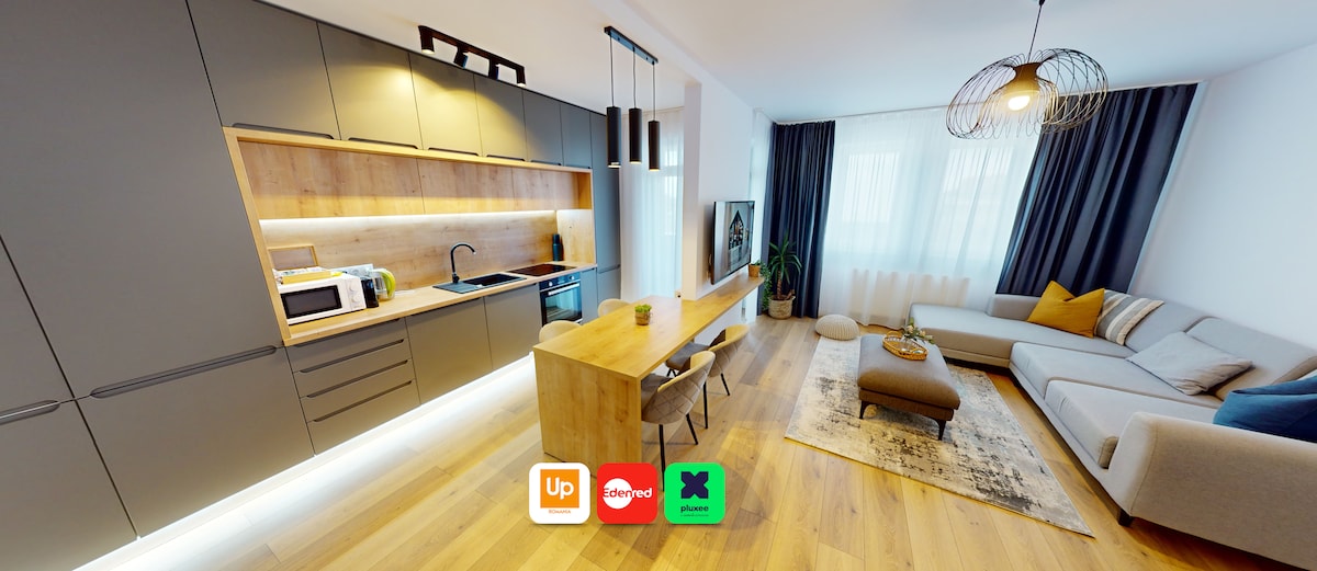 A modern open-space kitchen and living area features sleek cabinetry and a wooden dining table. Natural light filters through sheer curtains, illuminating cozy seating and decorative elements. The space combines functionality with comfort, providing a versatile atmosphere for relaxation or dining.