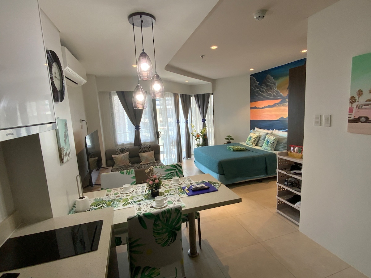 A modern studio apartment features an open layout with a kitchenette, dining area, and lounge space. Natural light filters through large windows, illuminating a cozy sofa and a spacious bed adorned in teal linens. A colorful mural enhances the backdrop, adding visual interest.