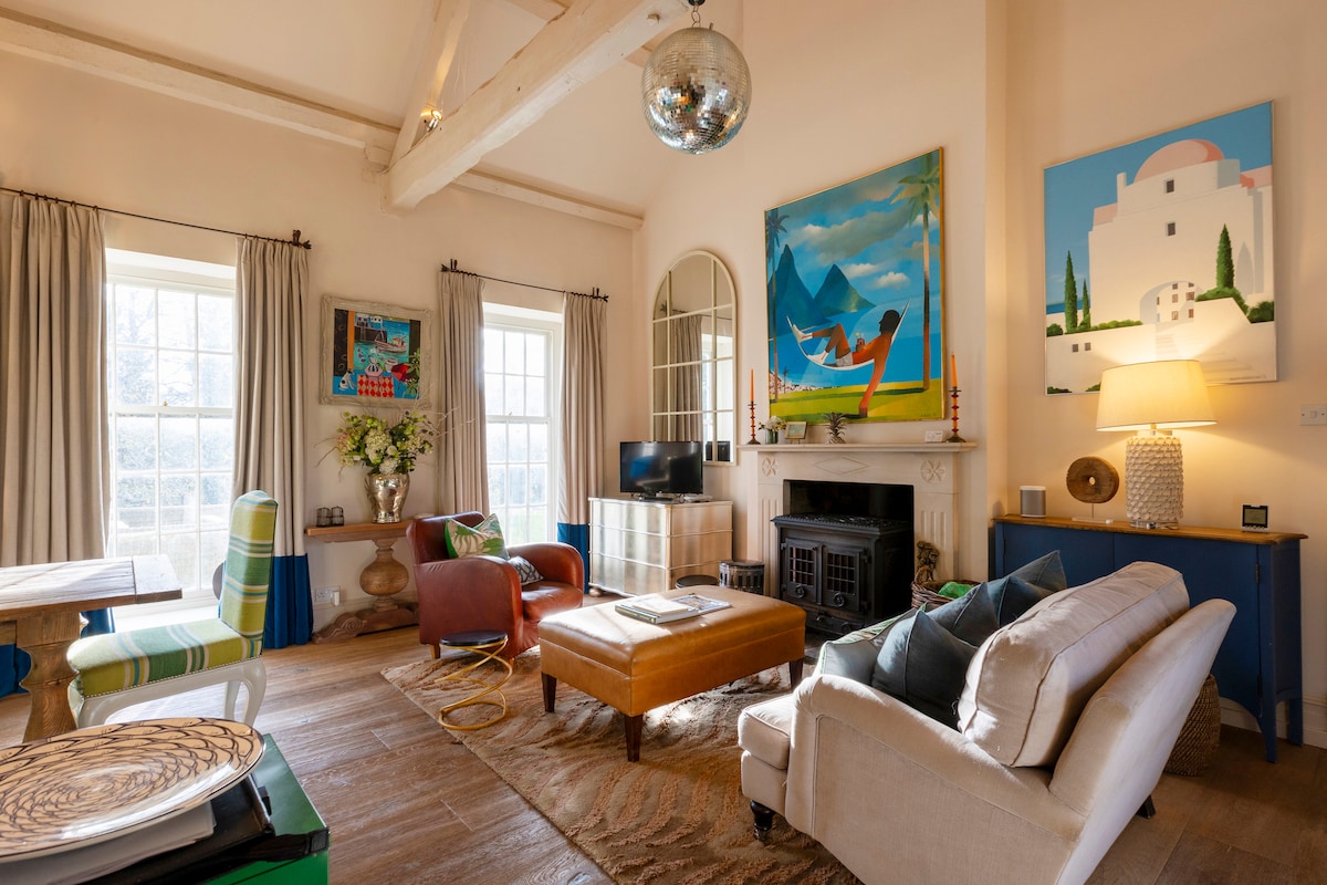A cozy sitting room features high ceilings with wooden beams and a large round mirror reflecting light. Colorful artwork adorns the walls, while an L-shaped sofa surrounds a coffee table. Natural light enters through large windows, and a tasteful lamp adds a soft glow.