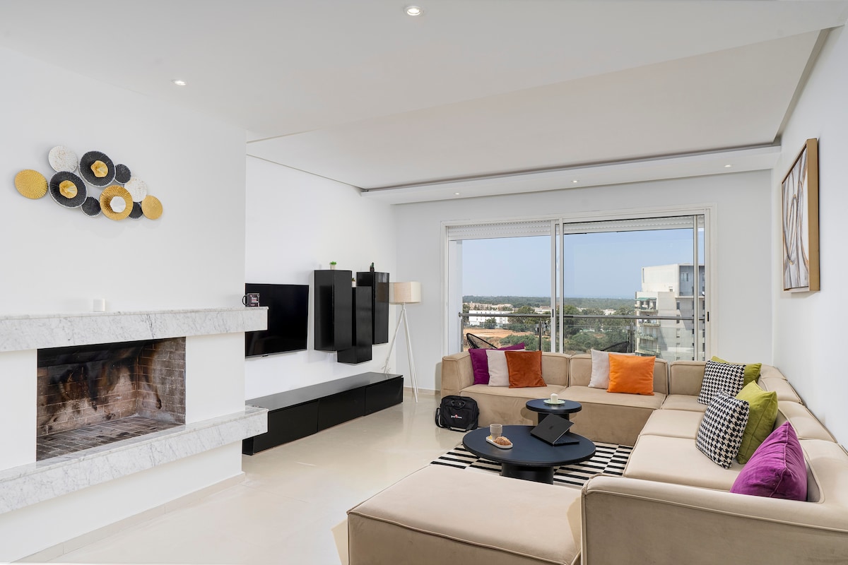 The living room features a spacious corner sofa adorned with colorful cushions. A modern coffee table sits on a light striped rug. Large windows provide a panoramic view of the surroundings, while a fireplace and wall-mounted television enhance the space's functionality.