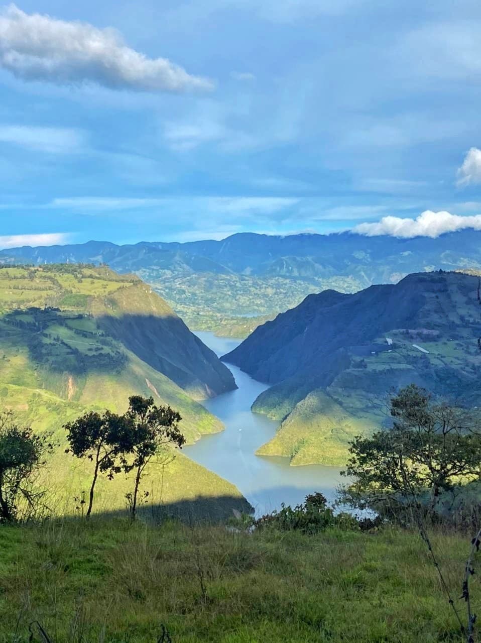 A panoramic view of the Embalse del Guavio showcases lush green mountains on either side, with the winding river reflecting the blue sky above. The landscape features varying shades of green and distant peaks, illustrating the natural beauty surrounding the area.