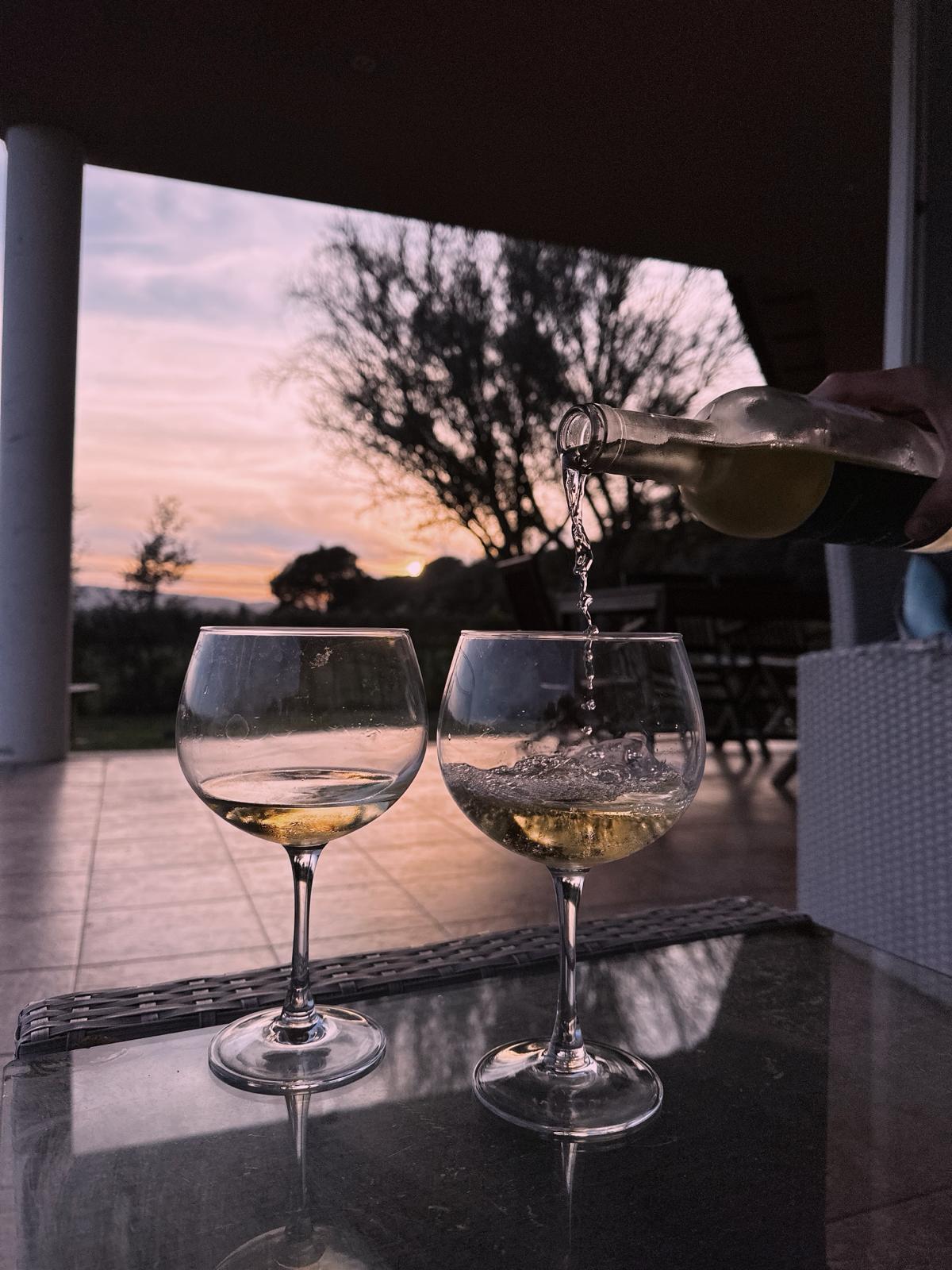 Two elegant glasses are filled with sparkling wine, set against a backdrop of a beautiful sunset. A hand is seen pouring wine from a bottle, while a blurred outdoor setting with greenery and furniture creates a relaxed atmosphere.