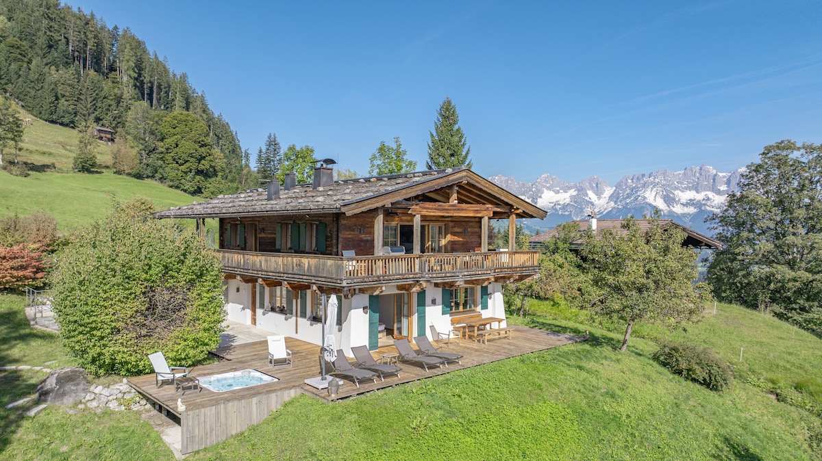 The chalet features a spacious outdoor terrace with sun loungers and a hot tub, set against a backdrop of lush greenery and majestic mountains. The wooden structure is complemented by stone accents and large windows, creating a seamless connection with the natural surroundings.