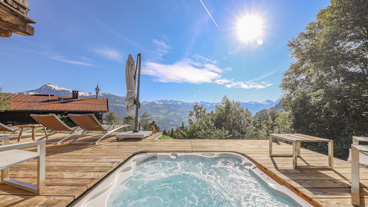 A private terrace features a hot tub surrounded by wooden decking, with sun loungers placed for relaxation. A mountain view is visible in the background, complemented by the clear blue sky and sunlight streaming down. A patio table adds to the outdoor space.