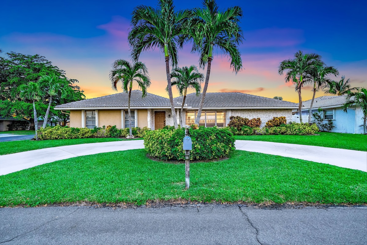 A well-maintained exterior of a single-story home is framed by lush green landscaping and palm trees. The driveway curves around a vibrant lawn, with a mailbox positioned at the front. A warm sunset provides a colorful backdrop to the serene setting.