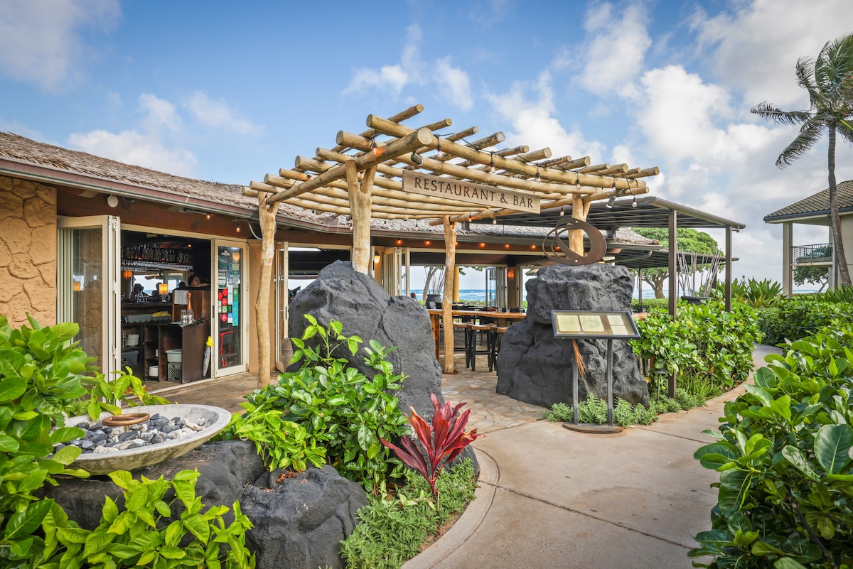 A beachside restaurant and bar entrance is framed by natural wooden beams and lush greenery. Smooth stone pathways lead to the entrance, where a sign denotes the establishment. The setting blends seamlessly into the tropical landscape with palm trees and a sky marked by soft clouds.