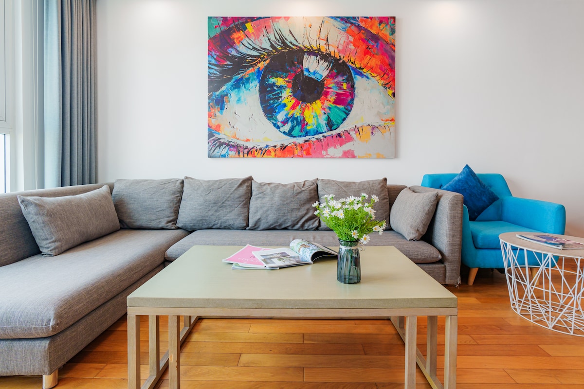 A modern lounge area features a spacious sectional sofa and a bright blue accent chair. A glass-top coffee table is topped with magazines and a small vase of fresh flowers, while a vibrant eye-themed artwork adds color to the white wall.