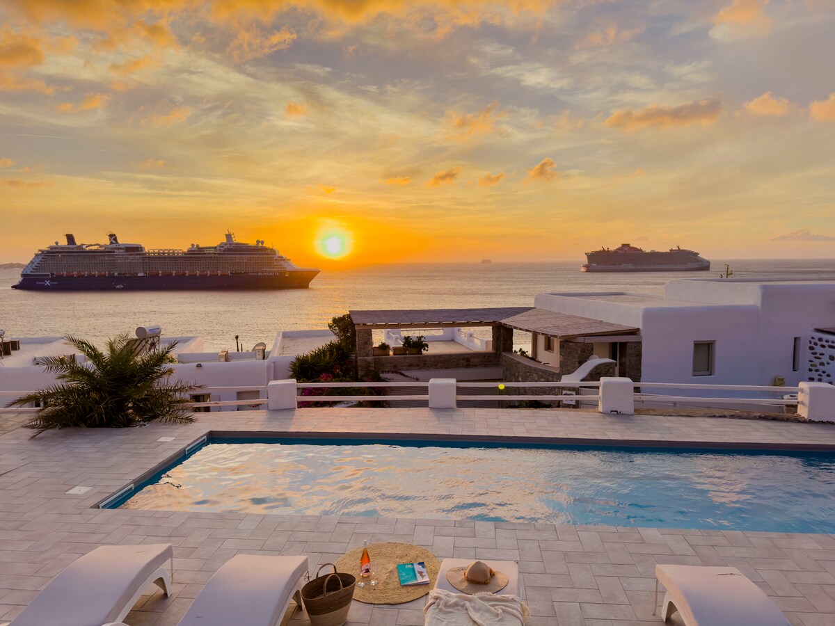 A serene pool area is shown with sun loungers positioned around a tranquil swimming pool. The vibrant sunset reflects off the water, while cruise ships are visible on the horizon, complementing the peaceful coastal landscape.