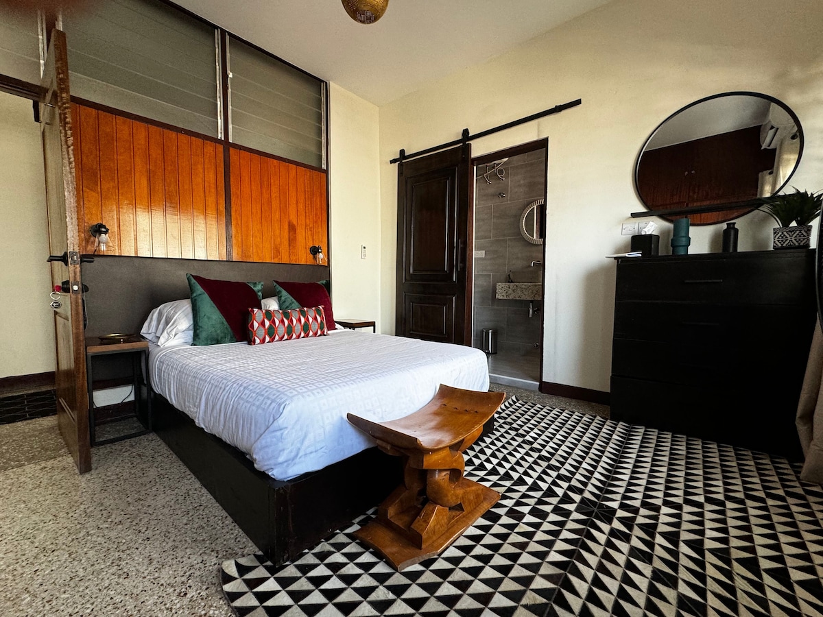 A modern bedroom features a spacious bed with decorative pillows against a backdrop of wooden accents. A patterned rug covers the floor, and a small wooden table adds character. Natural light enters through large windows, leading to an ensuite bathroom visible in the background.