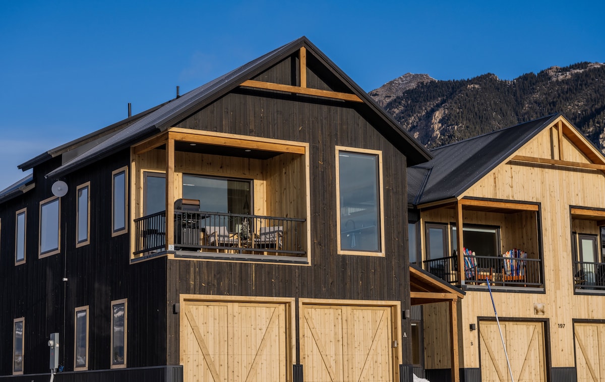 The exterior of a modern townhome is displayed, featuring a combination of dark wood and light wood paneling. The balcony is furnished with seating, offering a comfortable spot to enjoy the surrounding views. The mountain landscape serves as a backdrop.
