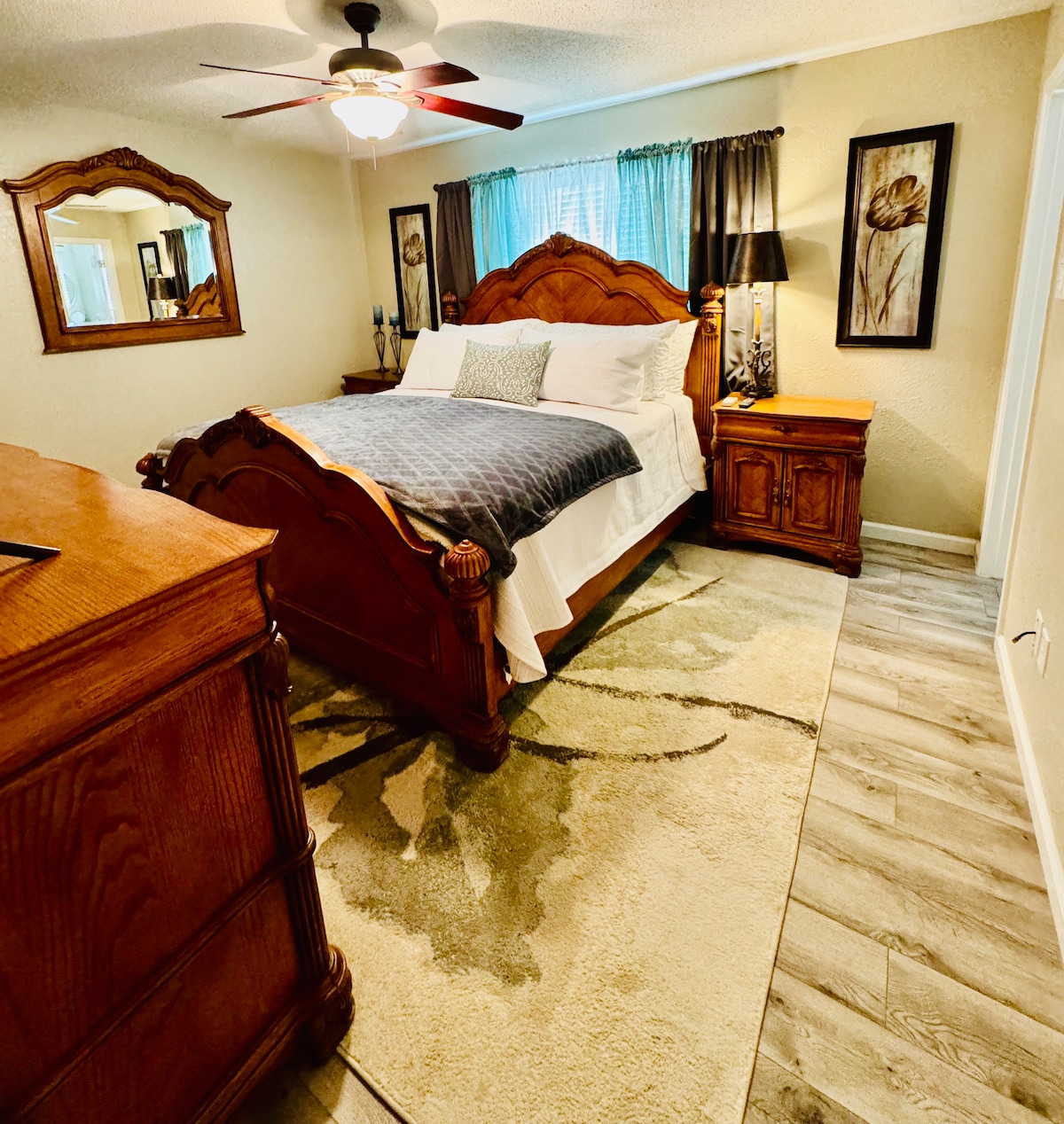 A well-furnished bedroom features a vaulted bed with a navy blanket, complemented by wooden nightstands and a dresser. Light is filtered through sheer curtains, creating a serene atmosphere. Artwork adorns the walls, while the floor showcases a soft area rug with floral accents.