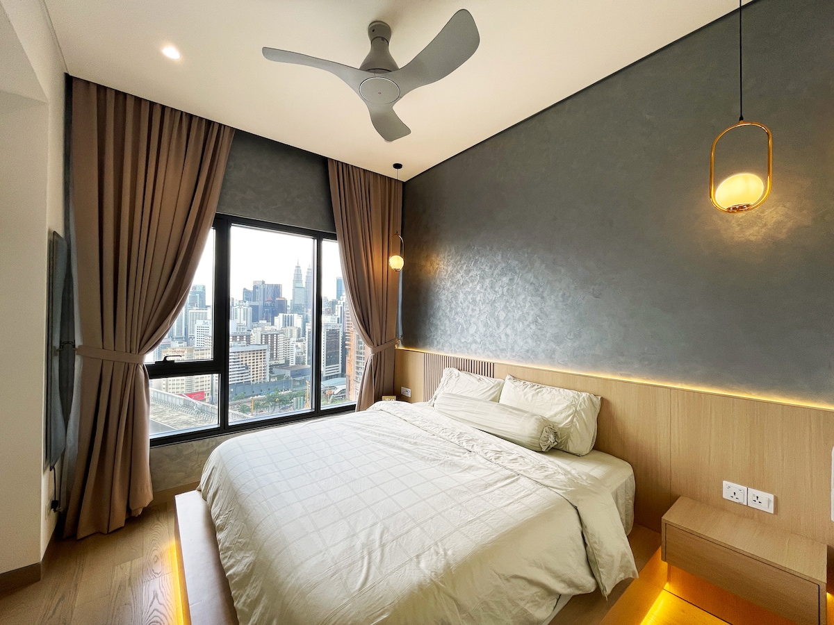 A spacious bedroom features a king-size bed with a light-colored duvet. Large windows showcase a city skyline view, while soft lighting from pendant fixtures creates a calming ambiance. Floor-to-ceiling curtains frame the windows, enhancing the modern decor.