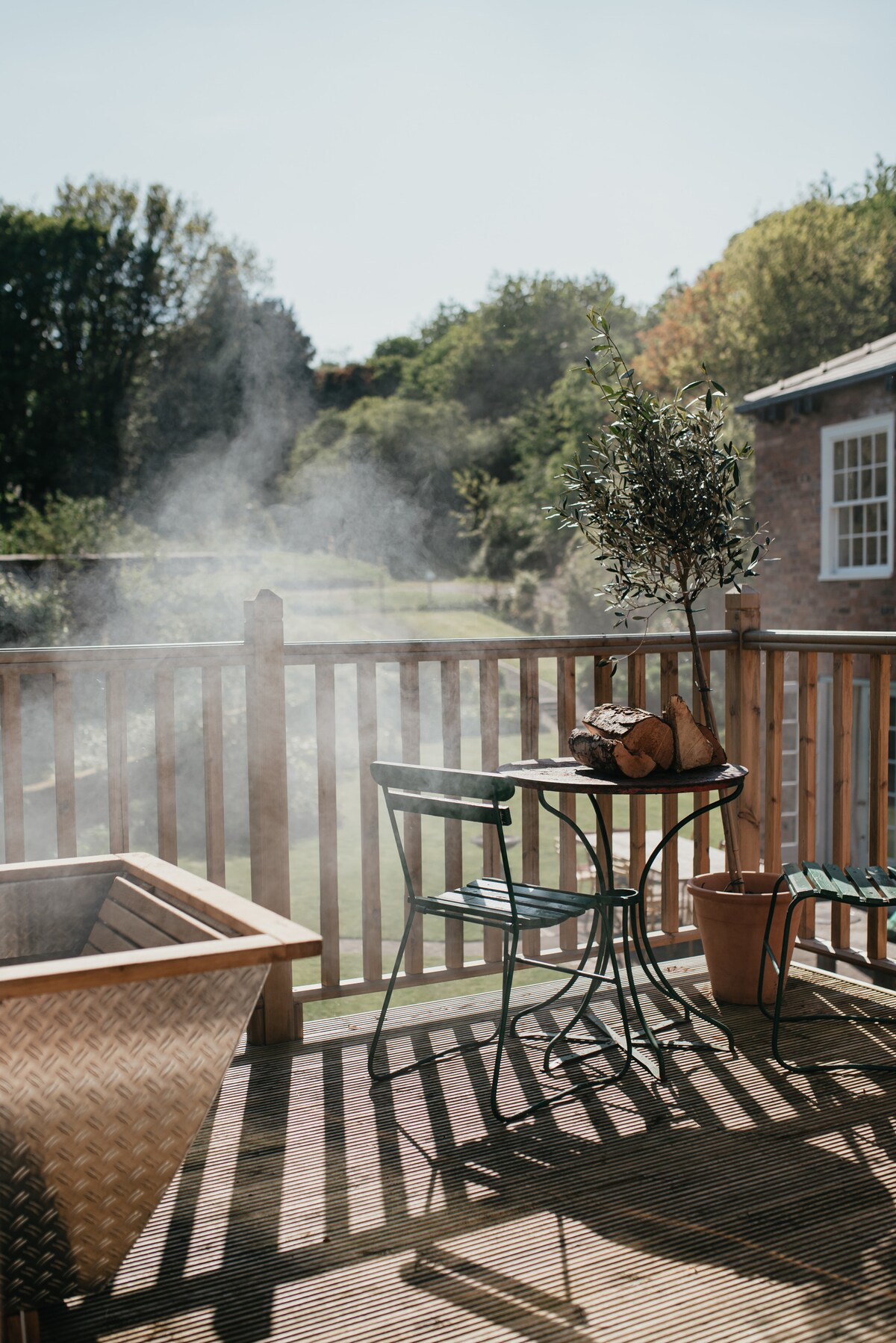 A wooden deck is seen, featuring a small circular table and two green metal chairs. A potted olive tree stands atop the table, and a wicker basket with pastries rests nearby. Light steam rises from a wood-fired hot tub situated adjacent to the deck.