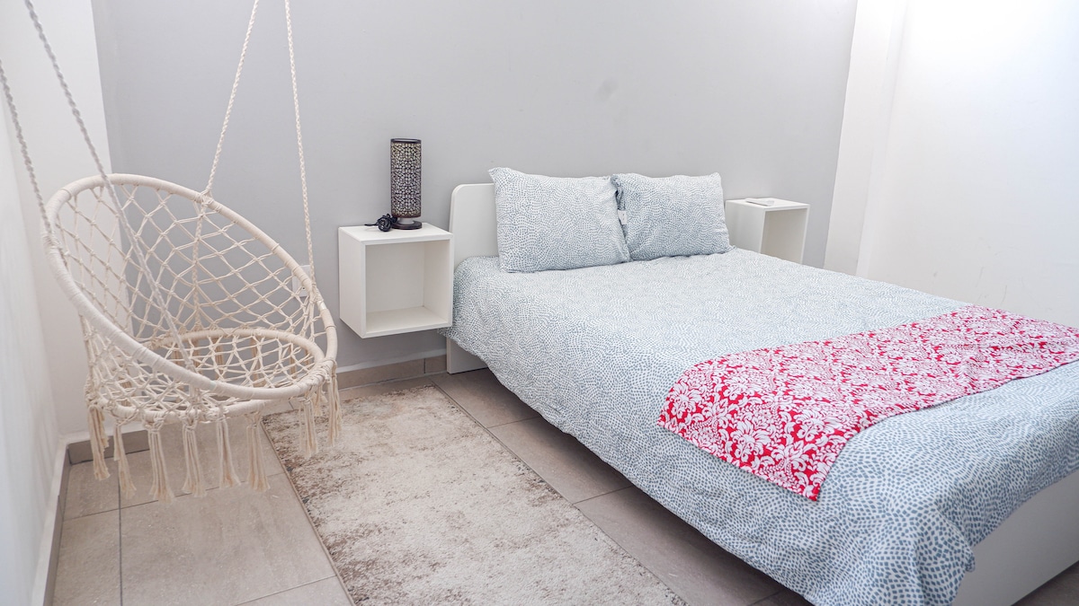 A calming sleeping area features a spacious bed dressed in light blue and white bedding, complemented by two bedside tables. A cozy hanging chair adds a unique touch. Soft natural light illuminates the room, highlighting the neutral color palette and tiled floor.