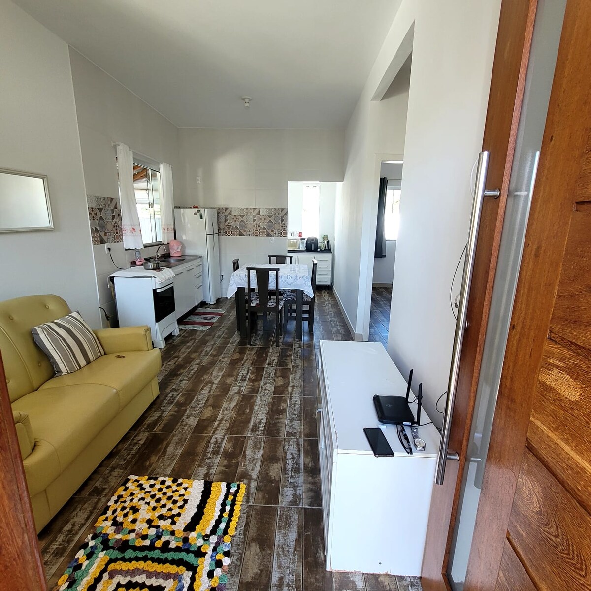 A bright and airy living space offers a welcoming view of an open kitchen and dining area. A cozy sofa is positioned next to a wooden dining table with several chairs. Natural light fills the room, enhancing the inviting atmosphere.