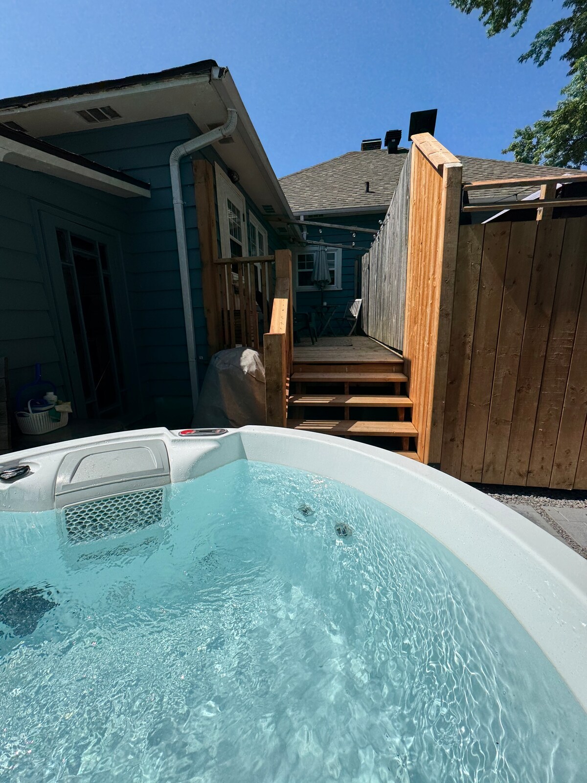 A hot tub with clear water is featured in the foreground, reflecting sunlight. In the background, wooden steps lead up to an enclosed area, bordered by fences. The surroundings include a section of a building with blue siding and a patio area visible beyond the steps.