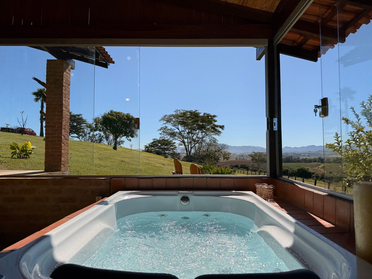 A spa with a jacuzzi is presented indoors, featuring clear views of the expansive green landscape and distant mountains through large glass windows. The calming blue water contrasts with the warm wood and brick elements of the space, creating an inviting focal point.