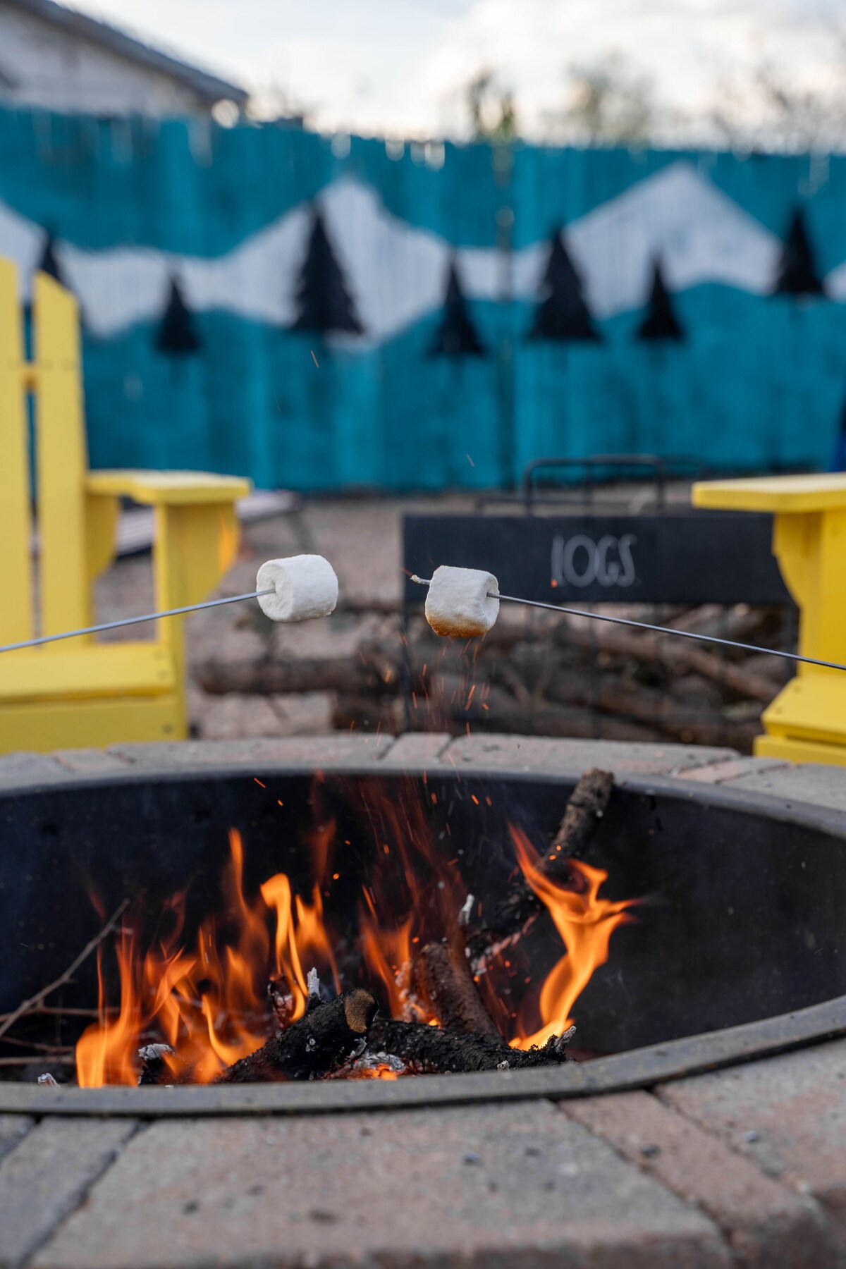 A fire pit is seen featuring vibrant flames, with marshmallows being roasted on sticks above. In the background, a blue and green painted fence displays silhouette trees. Bright yellow Adirondack chairs provide seating around the fire pit, creating an inviting outdoor atmosphere.