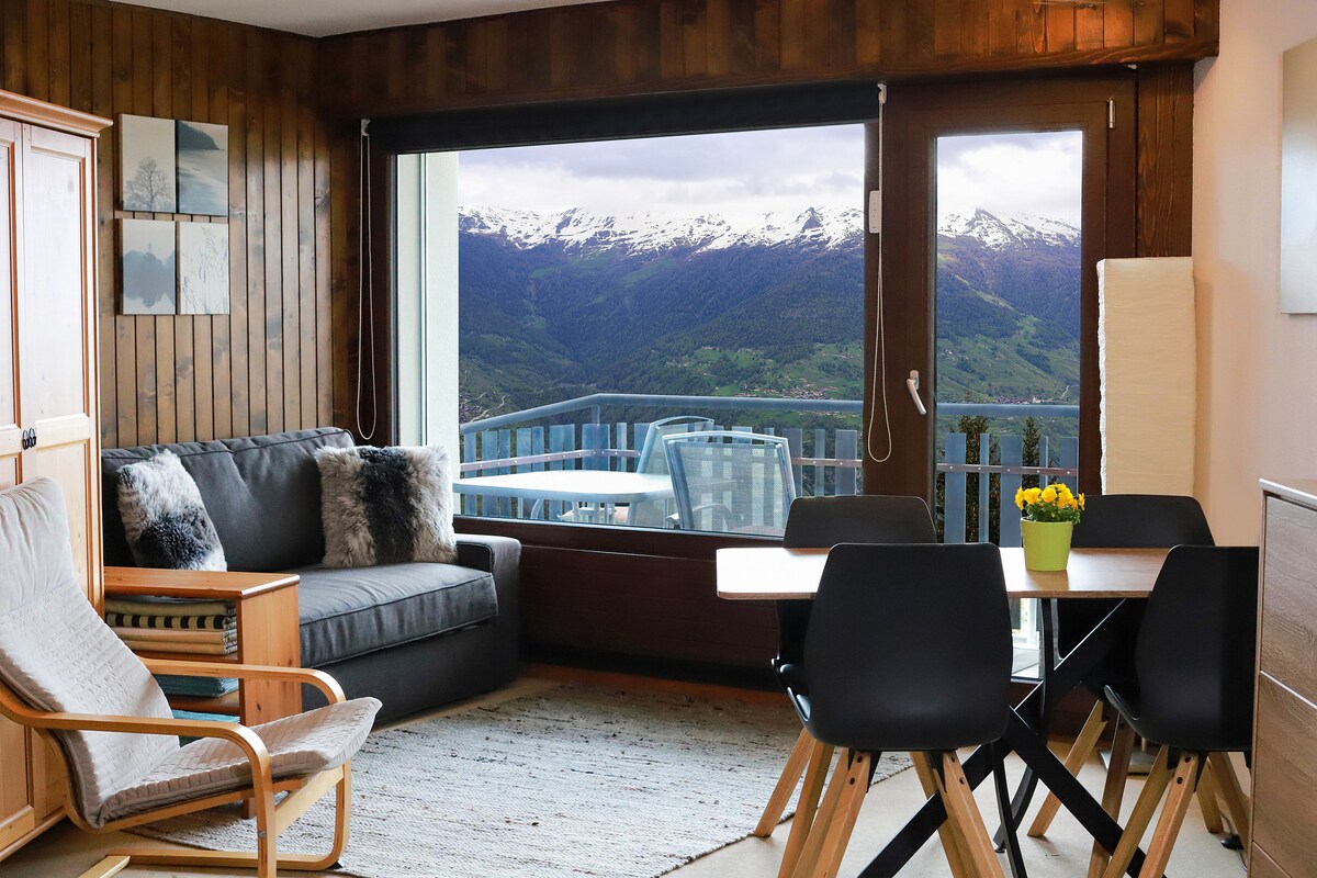 A spacious living area features a comfortable sofa and a lounge chair, complemented by wooden paneling. A dining table with black chairs is positioned nearby. Large windows offer expansive views of the mountains, while a balcony with outdoor seating can be seen in the distance.