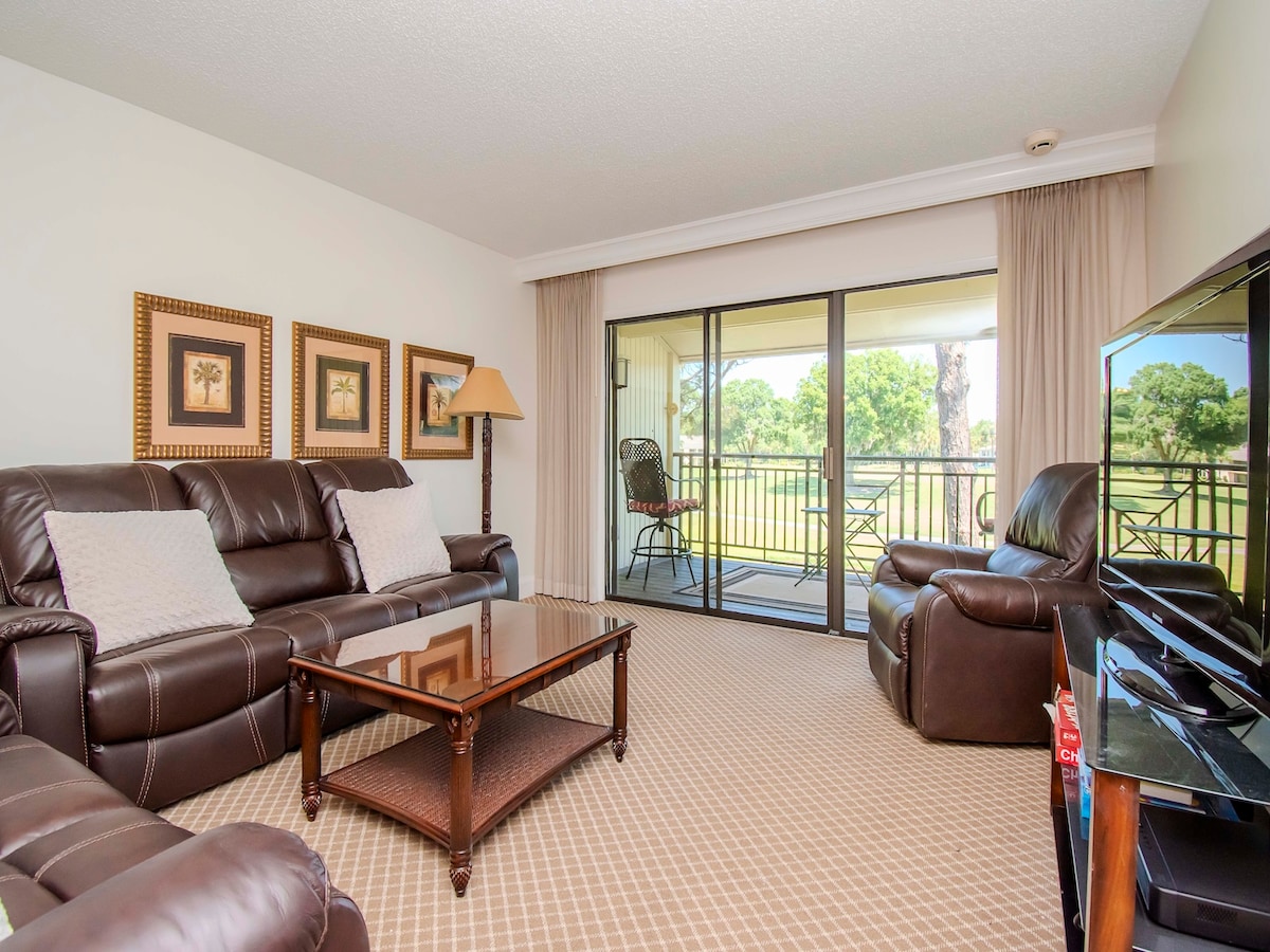 A cozy living area features a large glass coffee table at the center, surrounded by plush leather seating. Natural light is welcomed through sliding glass doors that lead to a patio, revealing scenic views of greenery beyond.